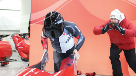 Bobsled competitor in helmet prepares to launch, coach signals beside.