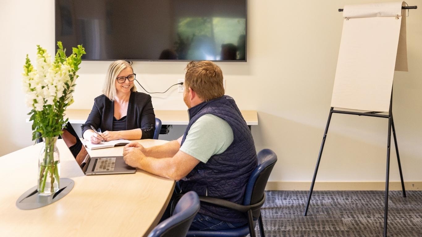 Two people talking at a table in a conference room with a flip chart.