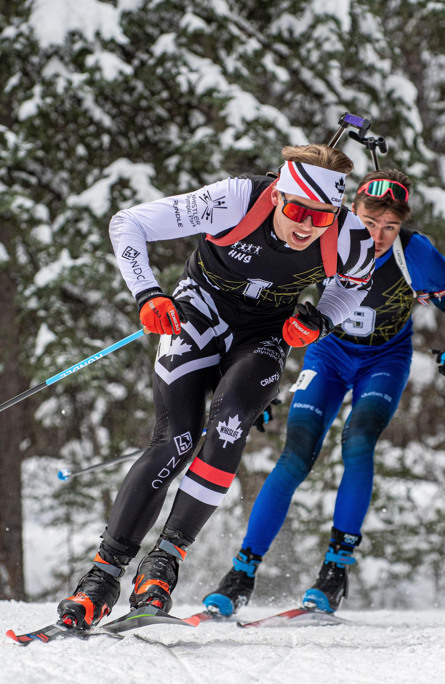 Skiers racing on snowy trail, wearing colorful gear, trees in background.