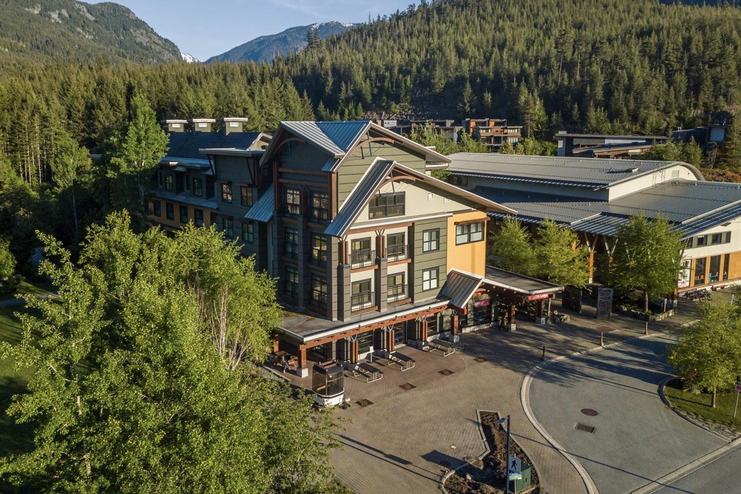 Large lodge with green siding surrounded by trees and mountains.
